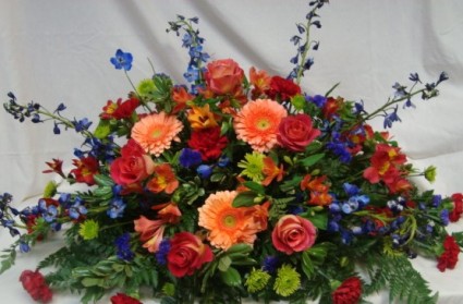 FOREVER TREASURED  Half Casket Spray of Seasonal shades of peach, reds, blues and lime greens. Roses, gerbera daisies, blue delphinium, button poms and more. 