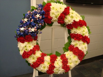 Red, White and Blue wreath with white Easel stand. Carnations, delphium and white cushion poms with ribbons and stars.
