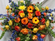 WARMEST REMEMBRANCE Half Casket Spray of seasonal shades of reds, oranges yellows and blues. Roses, gerbera daisies, delphinium, fugis and more.